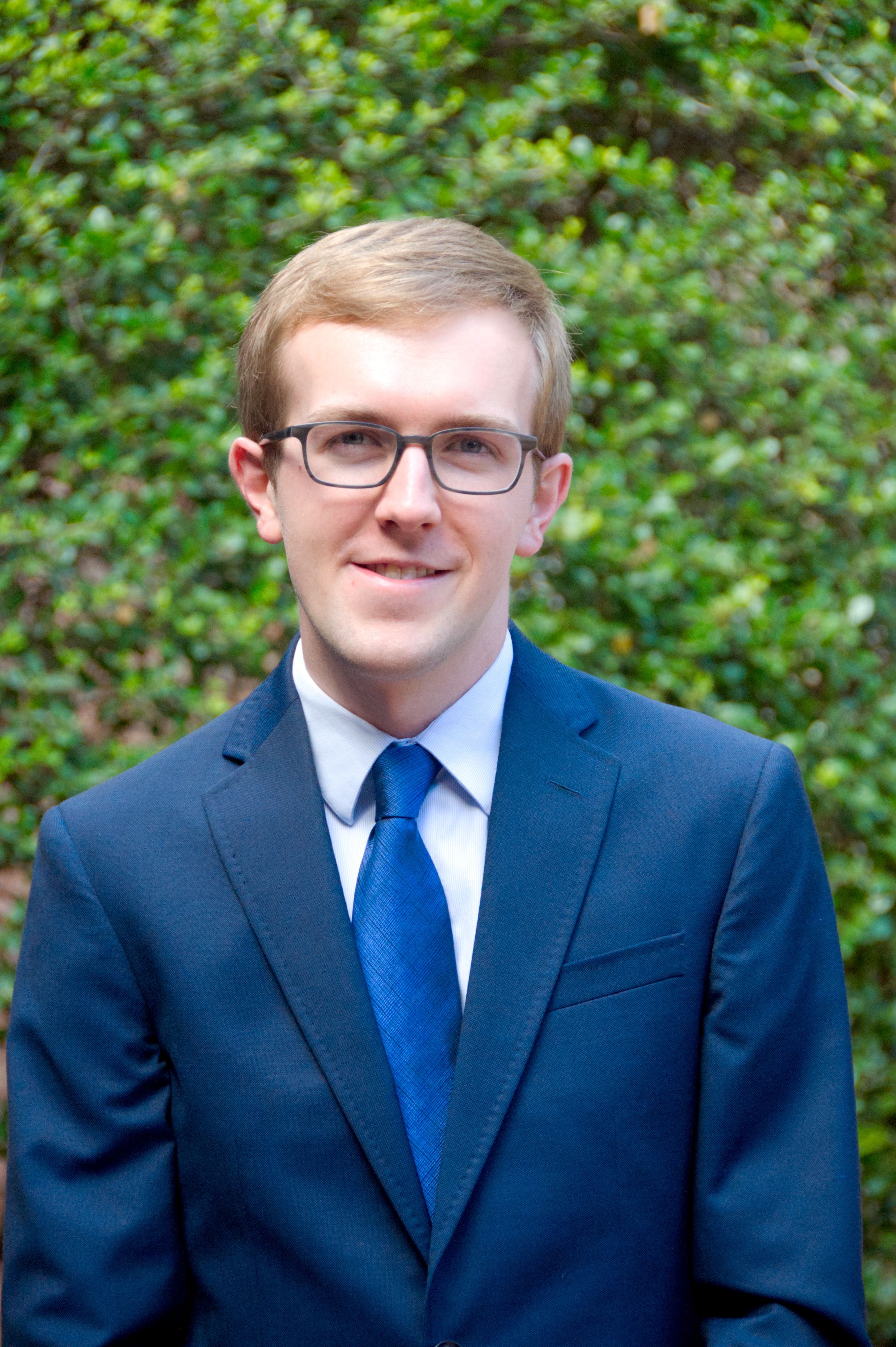 Jonathan in a blue suit and glasses in front of a green bush