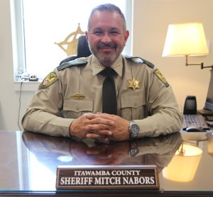 Photo of Sheriff Mitch Nabors smiling and sitting at a desk in front of a name plate